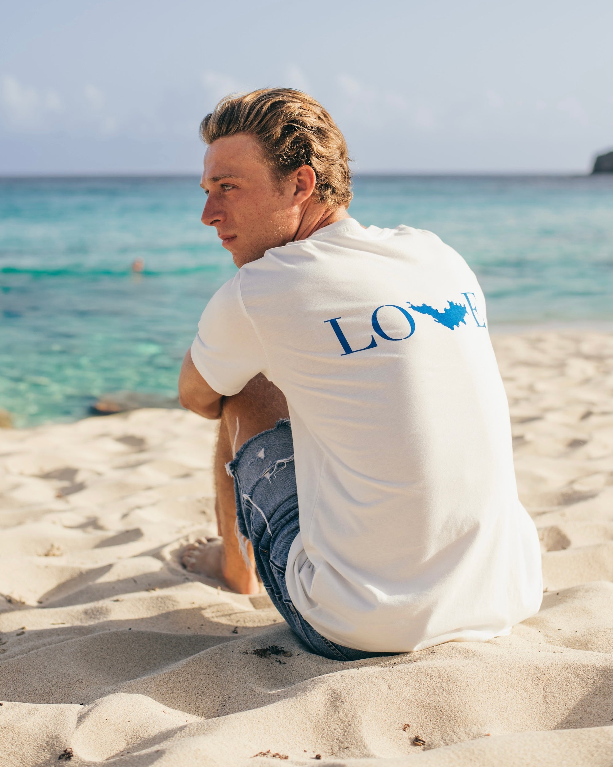 Martin porte le T-shirt Love St Barth blanc, assis dans le sable